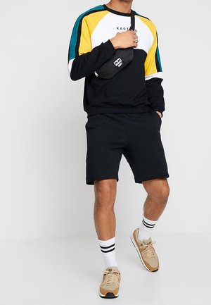 Hombre con sudadera de bloques de color, pantalones cortos negros, zapatillas beige, calcetines blancos con rayas y un bolso bandolera negro sobre un fondo blanco.