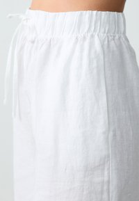 Pantalons en lin blanc avec une taille élastique, présentant un détail de cordon de serrage. Texture lisse et coupe ample. Coutures latérales visibles et tissu léger.