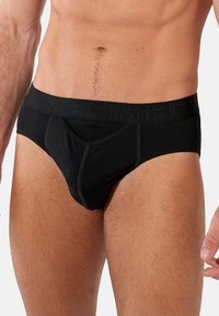 Schwarze Baumwollslips mit einem strukturierten Bund, der mit eingeprägtem Branding versehen ist. Das Design ist enganliegend mit einem konturierten Beutel und nahtlosen Kanten.