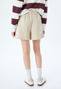Beige puuvillashortsit, joissa on joustava vyötärö, sileä pinta ja rento leikkaus. Päälle on yhdistetty valkoiset sukat ja vaaleat lenkkitossut.