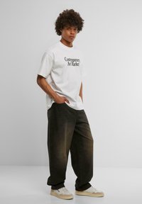 Hvit oversized t-skjorte med teksten "Contemporary Art Market", kombinert med mørke, smale bukser og beige lave sneakers. Glatt tekstur.