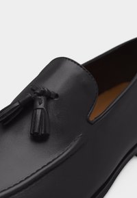 Mocassin en cuir noir avec bout arrondi, couture décorative et deux pampilles en cuir sur le dessus.