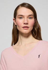 Ljust rosa v-ringad tröja i mjukt tyg, med en liten marinblå logotyp på vänster bröst och en avslappnad passform.