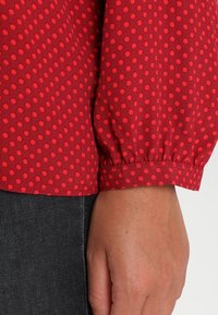 Rød bluse med prikker og lange, sammensnørede ærmer med manchet. Stoffet er glat og let, med jævnt store, kontrasterende prikker.