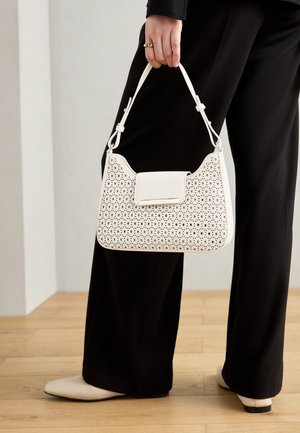 Persoon draagt zwarte broek en witte schoenen en houdt een witte geperforeerde handtas met een kort hengsel naast zich vast.