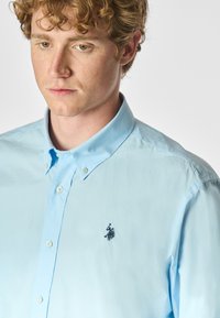 Camicia azzurra chiaro con colletto classico, dotata di un logo ricamato sul lato sinistro del petto e una texture di cotone morbido.