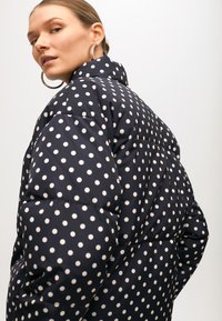 Námornická pufovaná bunda s bílými velkými puntíky, vysokým límcem a prošívanou texturou. Doplněná stříbrnými kruhovými náušnicemi.