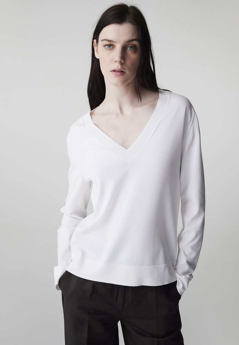 Joven con cabello oscuro y largo, vistiendo un suéter blanco con cuello en V y pantalones negros, de pie frente a un fondo claro y liso.