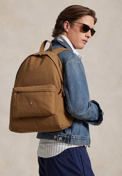 Άνδρας με γυαλιά ηλίου, τζην μπουφάν, ριγέ πουκάμισο και μπλε παντελόνι κουβαλάει ένα καφέ σακίδιο με ένα μικρό κεντημένο λογότυπο στην μπροστινή τσέπη.