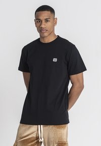 Camiseta de algodón negra con escote redondo y mangas cortas. Presenta un pequeño logotipo blanco en la parte izquierda del pecho. Diseño simple y clásico.