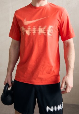 Personne portant un t-shirt Nike orange et un short Nike noir tenant un kettlebell noir dans la main droite, sur un fond uni.