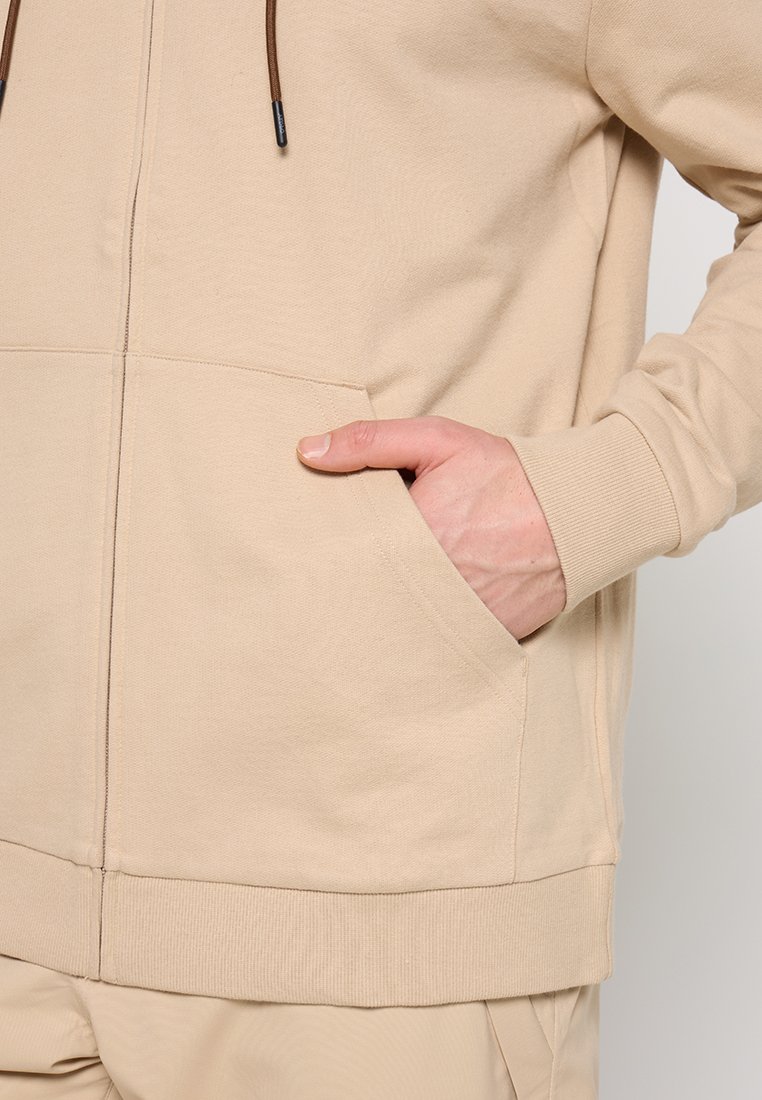 Hoodie beige zippé en tissu doux avec une poche kangourou et des cordons de serrage marron. Présente des poignets et un ourlet côtelés.