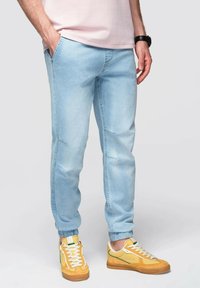 Světle modré džínové jogger kalhoty s manžetami na kotnících, s uvolněným střihem a jemnými švy, doplněné žlutými teniskami.