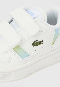 Valkoinen nahkainen lenkkari, jossa on tarrakiinnitys ja vihreä krokotiililogolla. Vaaleansinisiä yksityiskohtia sivuilla ja kuvioitu kumipohja.