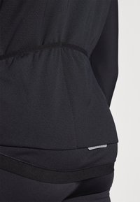 Black athletic top featuring a textured finish, gathered waist, and contrasting fabric at the hem. Includes a small logo accent at the back.