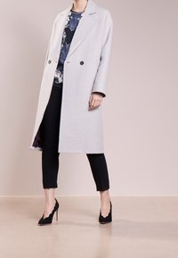 Abrigo gris claro de tejido texturizado, con un frente de doble botonadura, solapas muesca y bolsillos laterales, combinado con pantalones negros y tacones.