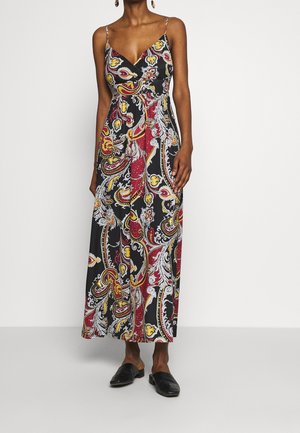 Kobieta ubrana w sukienkę bez rękawów sięgającą do kostek z wielokolorowym wzorem paisley, w czarnych wsuwanych butach i złotych kolczykach.