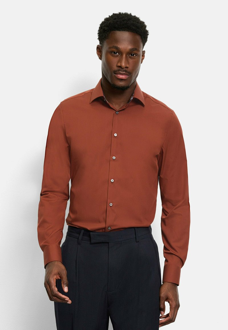 Rust-colored dress shirt with long sleeves, a button-down collar, and a subtle texture. Paired with dark trousers.