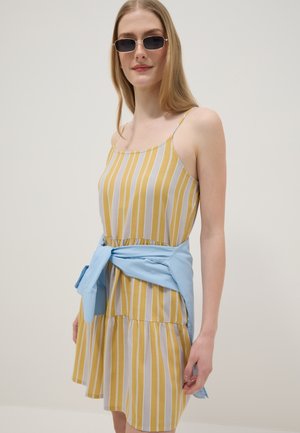 Woman wearing rectangular sunglasses and a yellow and white vertical striped sleeveless dress with a blue shirt tied around her waist.