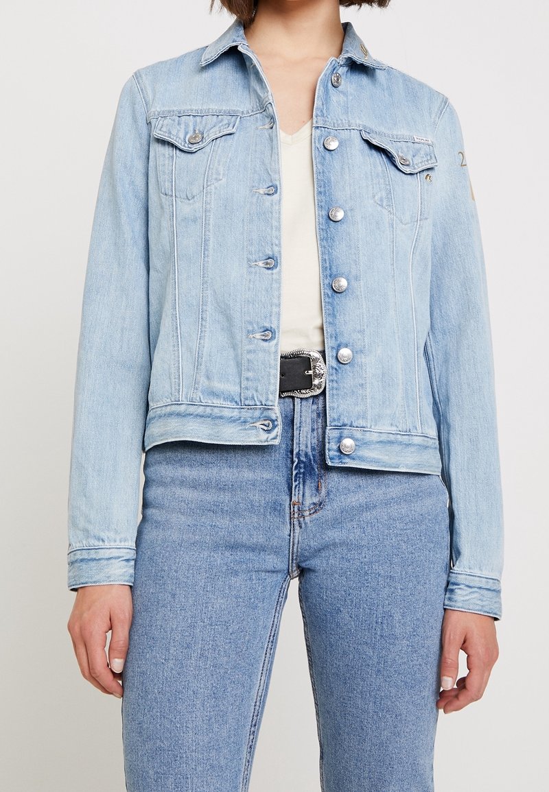 Light blue denim jacket with snap buttons, two chest pockets, and long sleeves. Worn over a white shirt and paired with blue jeans.