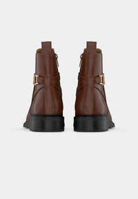 Paire de bottines en cuir marron avec semelles noires, fermeture éclair à l'arrière et boucles décoratives dorées sur les côtés.