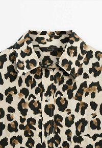 Chemise à imprimé léopard avec un fond beige orné de taches brun foncé et noires, col classique, boutonnière à l'avant, et détail d'une poche poitrine unique.