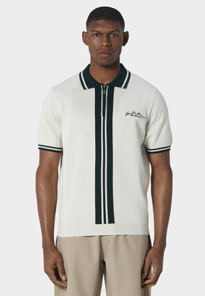 Man draagt een wit, kortärmelig gebreid poloshirt met zwarte dubbele strepen aan de voorkant en kraag, gecombineerd met beige broek.