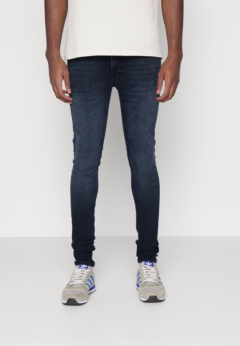 Vaqueros oscuros ajustados con un ligero desvanecimiento, fabricados en tela de mezclilla, combinados con zapatillas deportivas en gris y azul. Camiseta básica sin detalles.