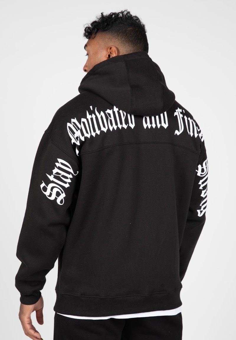 Melns kapučjak ar lielu balto tekstu uz muguras un piedurknēm. Iezīmē klasisku dizainu, plašu kapuci un ribotām piedurknēm.