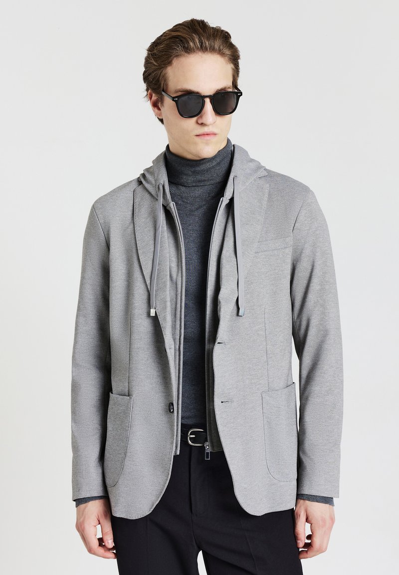 Giovane uomo che indossa occhiali da sole neri, giacca grigia a strati con cappuccio e tasche, dolcevita scuro e pantaloni neri su uno sfondo neutro.