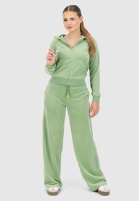 Le survêtement en velours vert comprend un hoodie zippé avec une poche avant et un pantalon ample avec un cordon de serrage réglable à la taille.