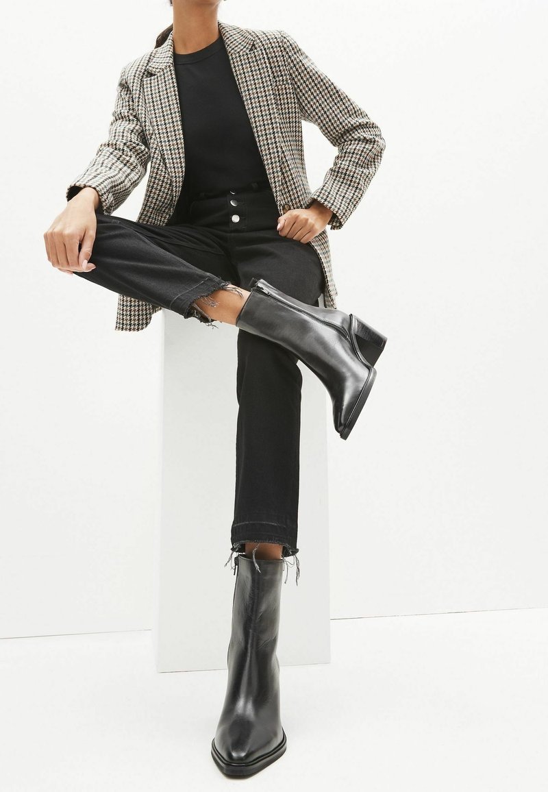 Personne portant des bottines noires à bouts pointus, un jean noir effiloché, un haut noir et un blazer à motif pied-de-poule, assise avec une jambe croisée sur l'autre.