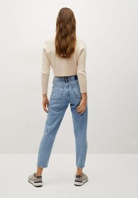 Suéter crema acanalado, vaqueros de talle alto azul claro, zapatillas grises. El cabello es largo y ondulado, con la vista trasera visible sobre un fondo neutro.