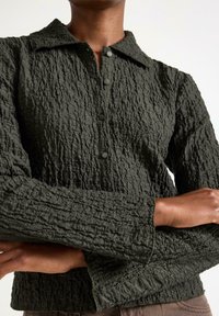 Mørkegrøn, struktureret skjorte med knapper og krave, der har et krøllet stofmønster, og lange ærmer med en let udvidet manchet.