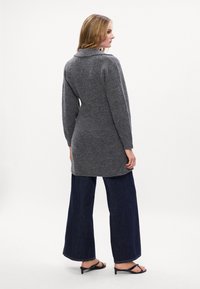 Μακρύ γκρι ζακέτα από μαλακό πλεκτό ύφασμα, ανοιχτό μπροστά, με γιακά σάλι. Συνδυασμένη με φαρδιά σκουρόχρωμα τζιν παντελόνια και μαύρα σανδάλια.