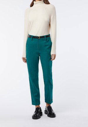 Türkise Hose mit geradem Schnitt, mit umgeschlagenen Säumen und einem Gürtel; kombiniert mit einem cremefarbenen Rollkragenpullover. Schwarze Schnürschuhe runden das Outfit ab.
