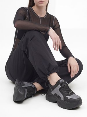 Persona sentada con las piernas cruzadas, vistiendo una blusa negra transparente, pantalones cargo negros y zapatillas gruesas negras y grises.