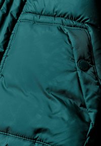 Teal puffer jakna s kviltanom teksturom. Sadrži bočnu džep s kružnim dugmetom za zatvaranje. Glatka završna obrada materijala.