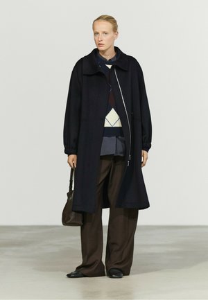 Femme portant un long manteau noir par-dessus un pull à motifs et un pantalon large marron, tenant un sac à main marron foncé, debout sur un sol clair.