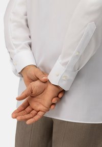 Witte langarmige shirt van soepele stof, met schuine manchetten en ronde, witte knopen. Handen op de rug geklapt, in beige broek.