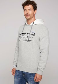 Felpa grigia realizzata in tessuto morbido, con una fodera bianca, cordoncini e il testo "Camp David Flight Crew" in colori e caratteri variabili.