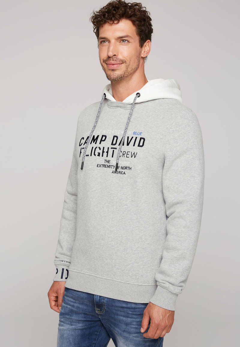 Felpa grigia realizzata in tessuto morbido, con una fodera bianca, cordoncini e il testo "Camp David Flight Crew" in colori e caratteri variabili.