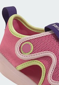 Ružová sieťovaná topánka s fialovým pásikom, zelenými akcentmi a textúrovaným stehom. Obsahuje detail s okrúlym gombíkom a otvorené výrezy pre lepšiu priedušnosť.