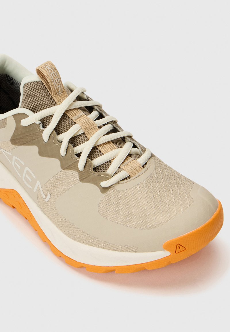 Zapatilla deportiva beige con parte superior de malla, suela de goma, acentos naranjas y cordones blancos; presenta una superficie texturizada y una puntera reforzada.
