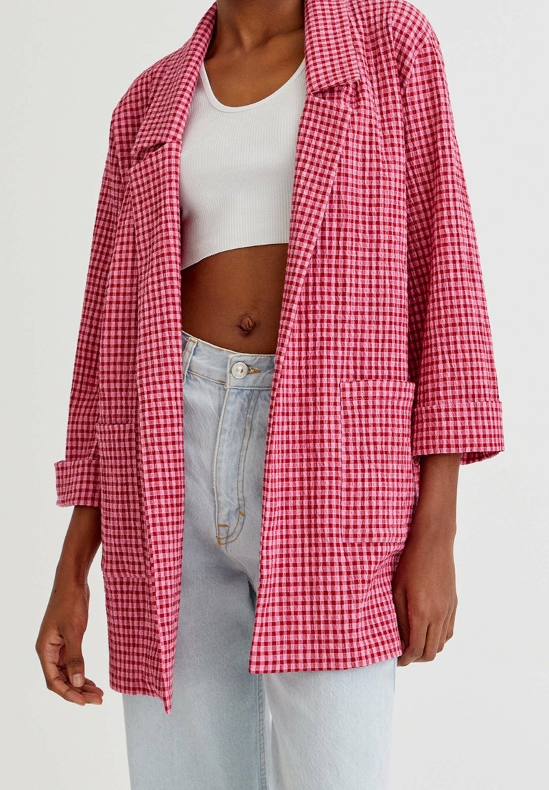Femme portant un jean clair, un débardeur court blanc et un blazer ample à carreaux roses et rouges avec les manches retroussées.
