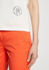 Bílé tričko s krátkým rukávem s kulatým logem a textem "Tommy Hilfiger"; spárované s jasně oranžovými kalhotami s hladkou texturou.