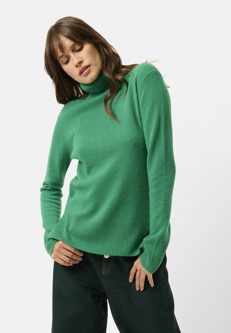 Maglione verde a collo alto in un morbido knit testurizzato con polsini e orlo a coste, abbinato a pantaloni scuri per creare contrasto.