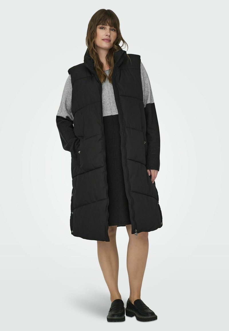 Svart stoppad lång väst med hög krage, framdragkedja, sidofickor och en slät textur, lager på lager över en grå tröja och kjol.