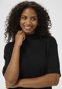 Zwart top met korte mouwen en hoge hals, gemaakt van een textuurstof met subtiele glitters, gedragen door een glimlachend persoon die zijn of haar kin aanraakt.