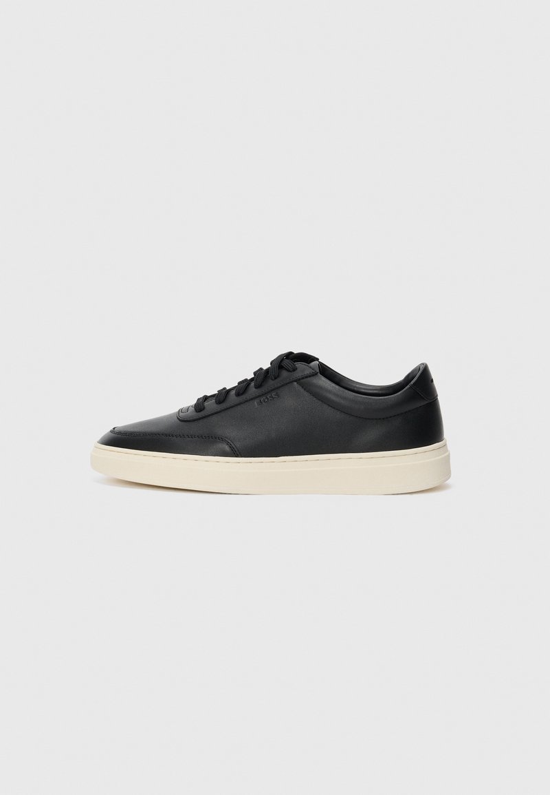 Mustad musta nahast madal sneaker mustade nööpidega ja kreemivalge tallaga, nähtud külgprofiilis valge tausta ees.